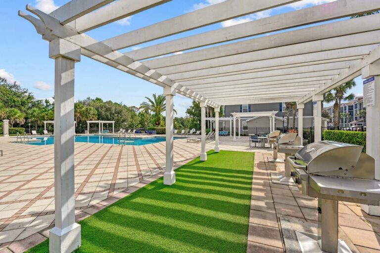 Outdoor grill area under a stylish pergola, vibrant green landscaping, and a swimming pool area in a serene setting.