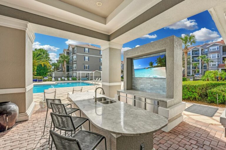 Outdoor kitchen with sleek countertops overlooking a swimming pool, framed by lush greenery and stylish apartment buildings, perfect for entertaining.